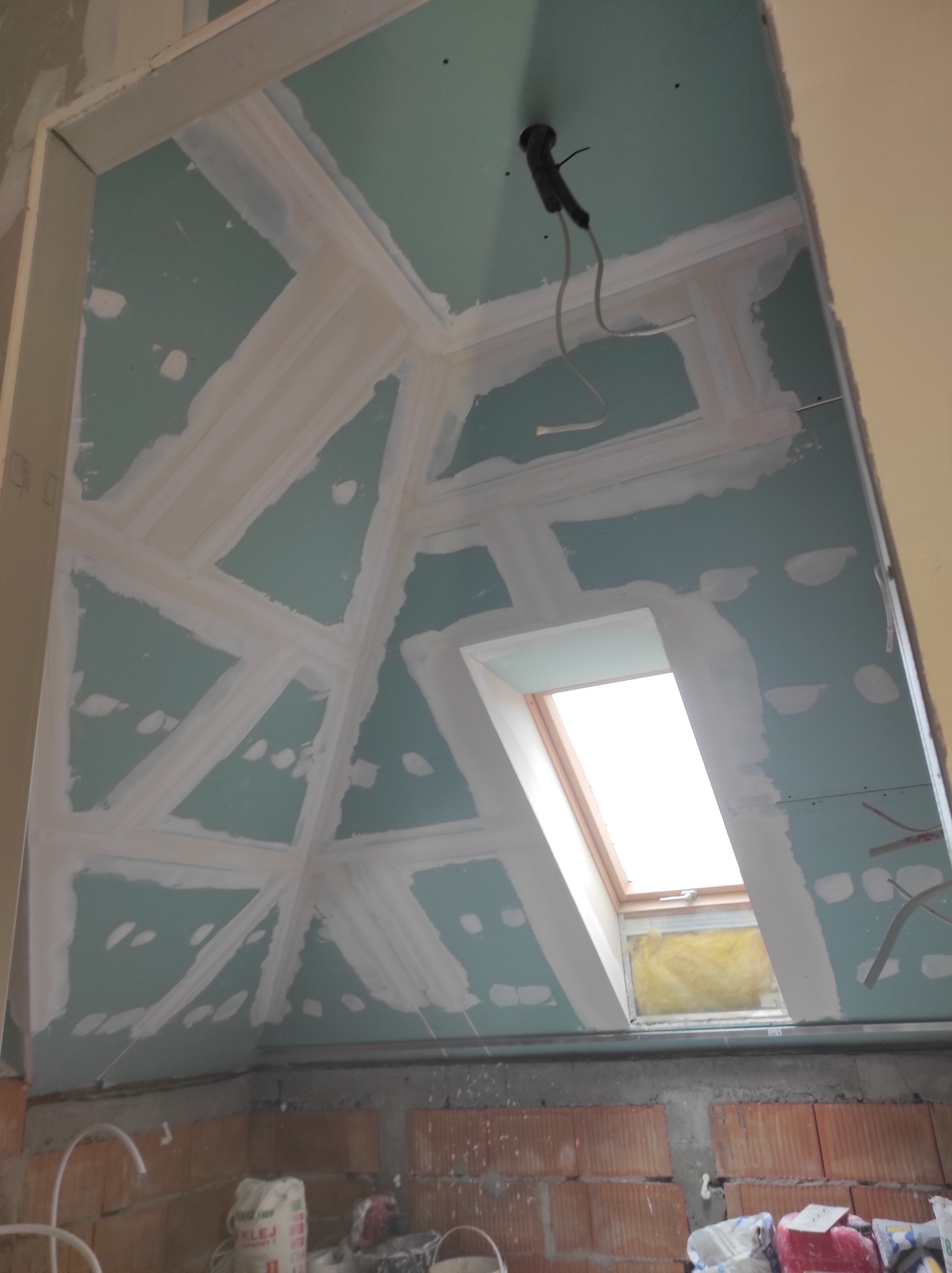 Skośny sufit poddasza z oknem dachowym w trakcie zabudowy płytami gipsowo-kartonowymi i szpachlowania łączeń, widoczne kable elektryczne i ceglana ściana w tle.