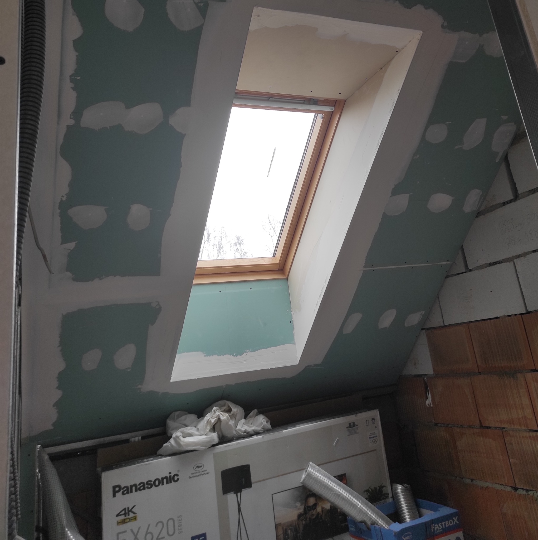 Montaż okna dachowego w skośnej ścianie, widoczne płyty gipsowo-kartonowe z nałożoną gładzią szpachlową, drewniana rama okienna i kartony na podłodze.