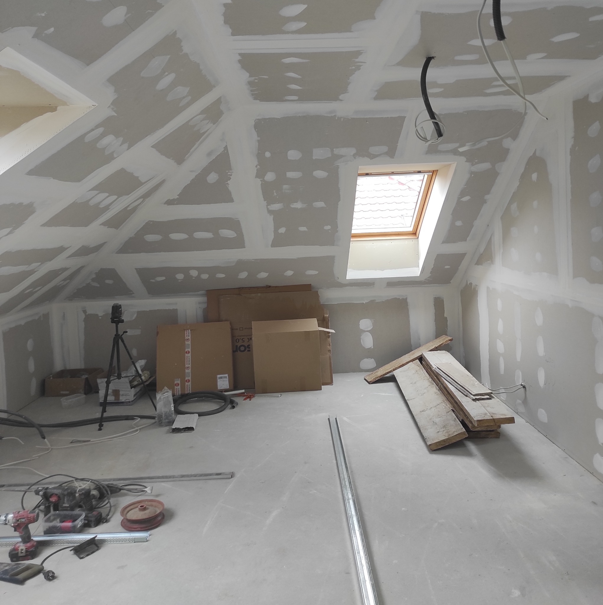 Poddasze w trakcie adaptacji: widoczne płyty gipsowo-kartonowe na skośnym suficie i ścianach, częściowo zaszpachlowane, okno dachowe, narzędzia oraz materiały budowlane na podłodze.