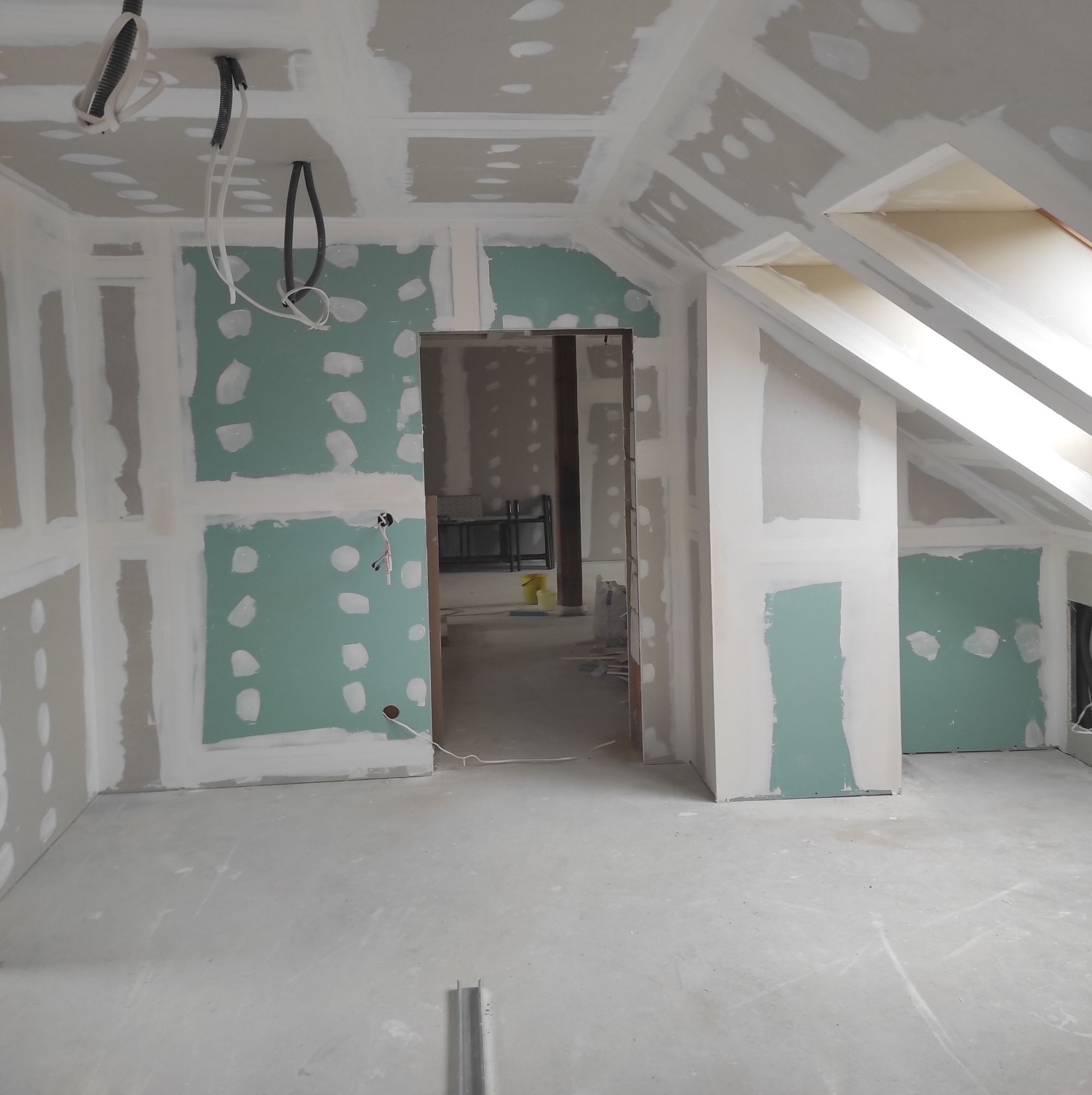 Pokój na poddaszu w trakcie montażu płyt gipsowo-kartonowych na ścianach i suficie, widoczne kable elektryczne wystające z sufitu, szpachlowane łączenia płyt i otwór drzwiowy do kolejnego...