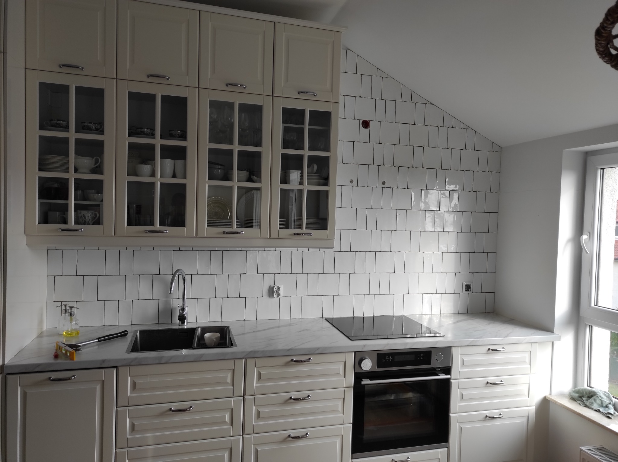 Jasna kuchnia w stylu klasycznym z białymi, prostokątnymi płytkami na ścianie, marmurowym blatem i przeszklonymi szafkami wypełnionymi naczyniami.