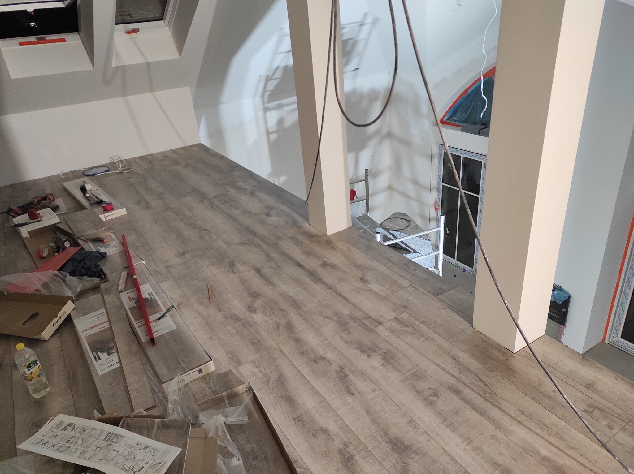 Montaż paneli podłogowych w jasnym, przestronnym poddaszu z widocznymi narzędziami i materiałami budowlanymi KronoStep. W tle widoczne kolumny, okno dachowe i schody.