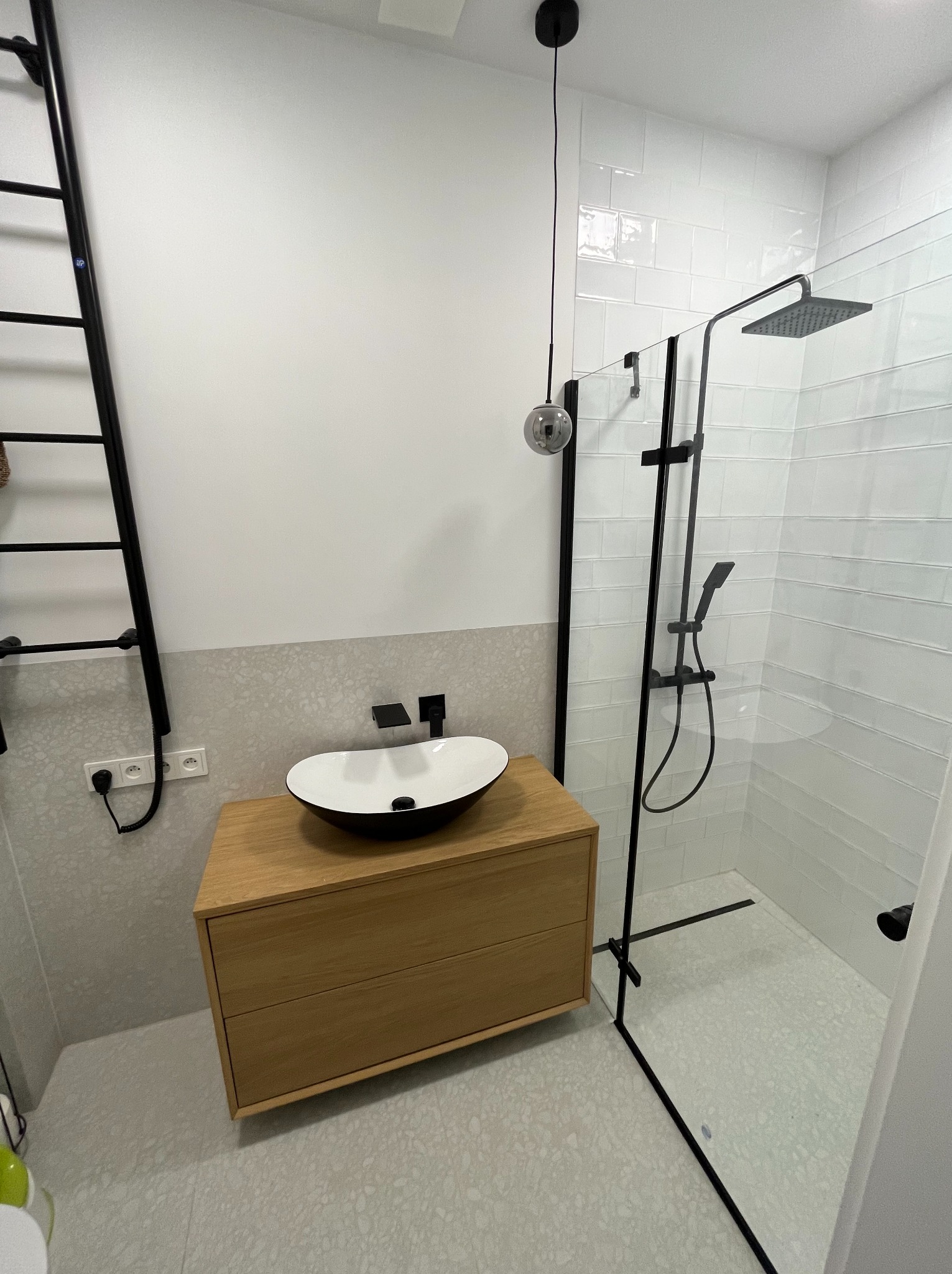 Nowoczesna łazienka z kabiną prysznicową typu walk-in z czarnymi detalami, wisząca szafka z umywalką nablatową o owalnym kształcie, białe płytki ułożone w cegiełkę, dekoracyjny grzejnik drabinkowy...