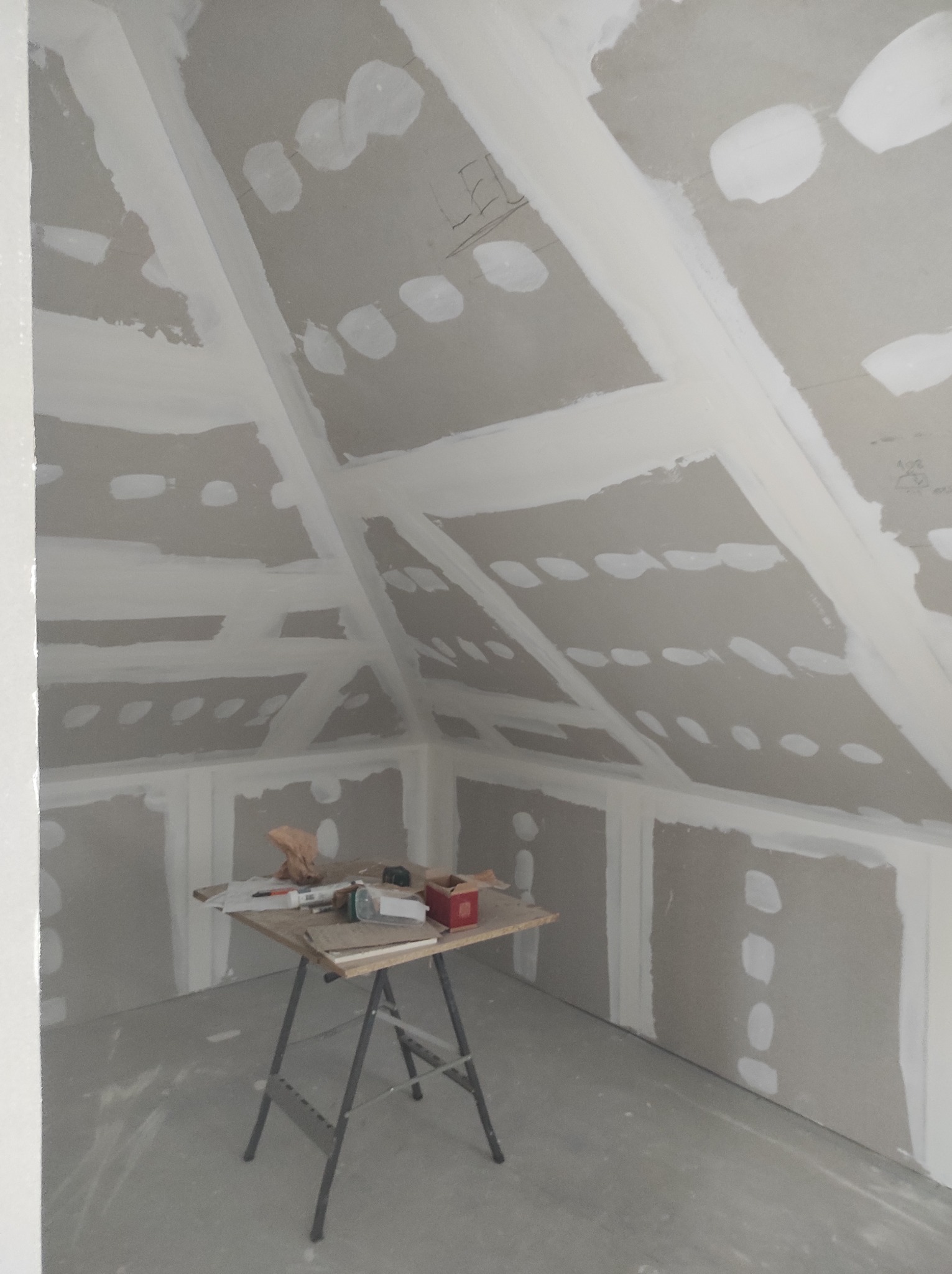 Poddasze w trakcie zabudowy płytami gipsowo-kartonowymi; widoczne szpachlowane łączenia, stół roboczy z narzędziami i materiałami budowlanymi w niezagospodarowanym pomieszczeniu.