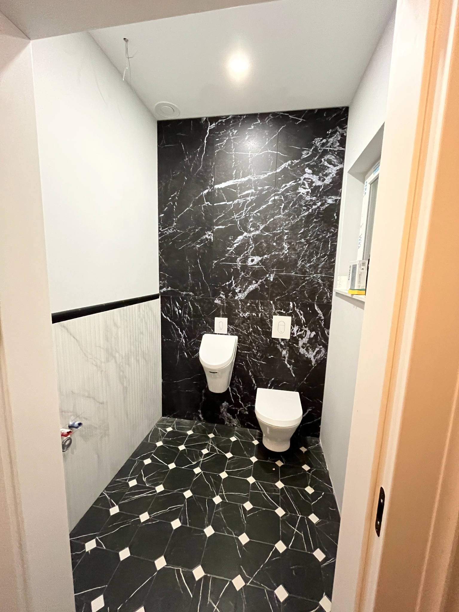 Wykończona łazienka z czarno-białymi płytkami imitującymi marmur na ścianach i podłodze, wisząca toaleta i bidet, białe ściany częściowo wykończone pionowymi panelami.