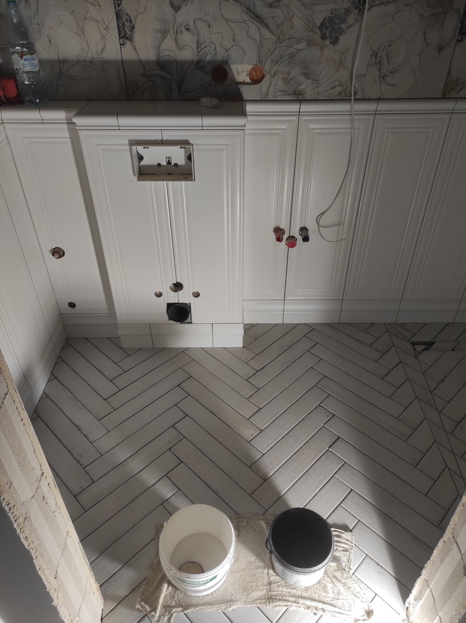 Wnętrze łazienki w trakcie prac wykończeniowych: białe płytki ułożone w jodełkę na podłodze, białe szafki z otworami na instalacje, widoczne dwa wiadra i fragment ściany przed wykończeniem.