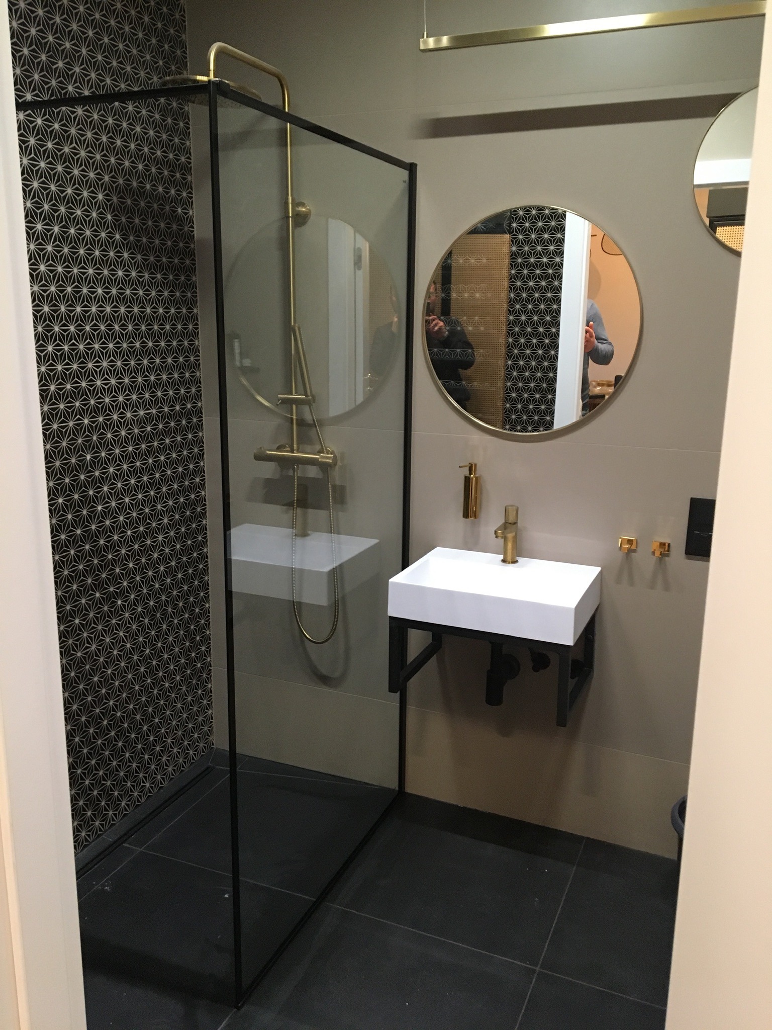 Nowoczesna łazienka z kabiną prysznicową typu walk-in z czarną ramą, geometryczną mozaiką na ścianie, białą umywalką na czarnym stelażu i okrągłym lustrem z złotą ramą, całość w stonowanej...