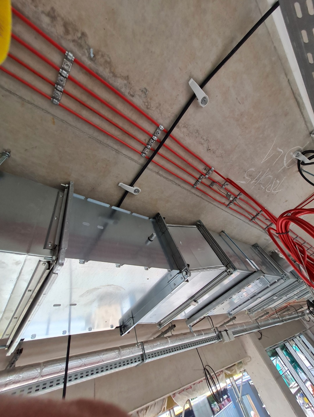 Widok z dołu na sufit z zamontowanymi metalowymi korytkami kablowymi i czerwonymi przewodami w peszlach, przymocowanymi klamrami do sufitu, widoczne także uchwyty do przewodów.