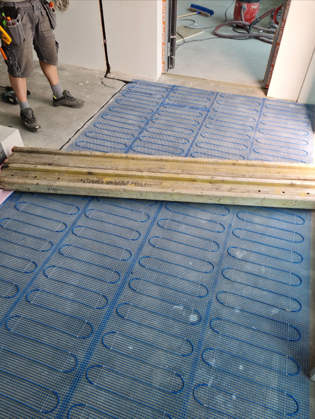 Instalacja maty grzewczej podłogowej wewnątrz pomieszczenia, widoczne elementy instalacji i fragment roboczego stroju instalatora.