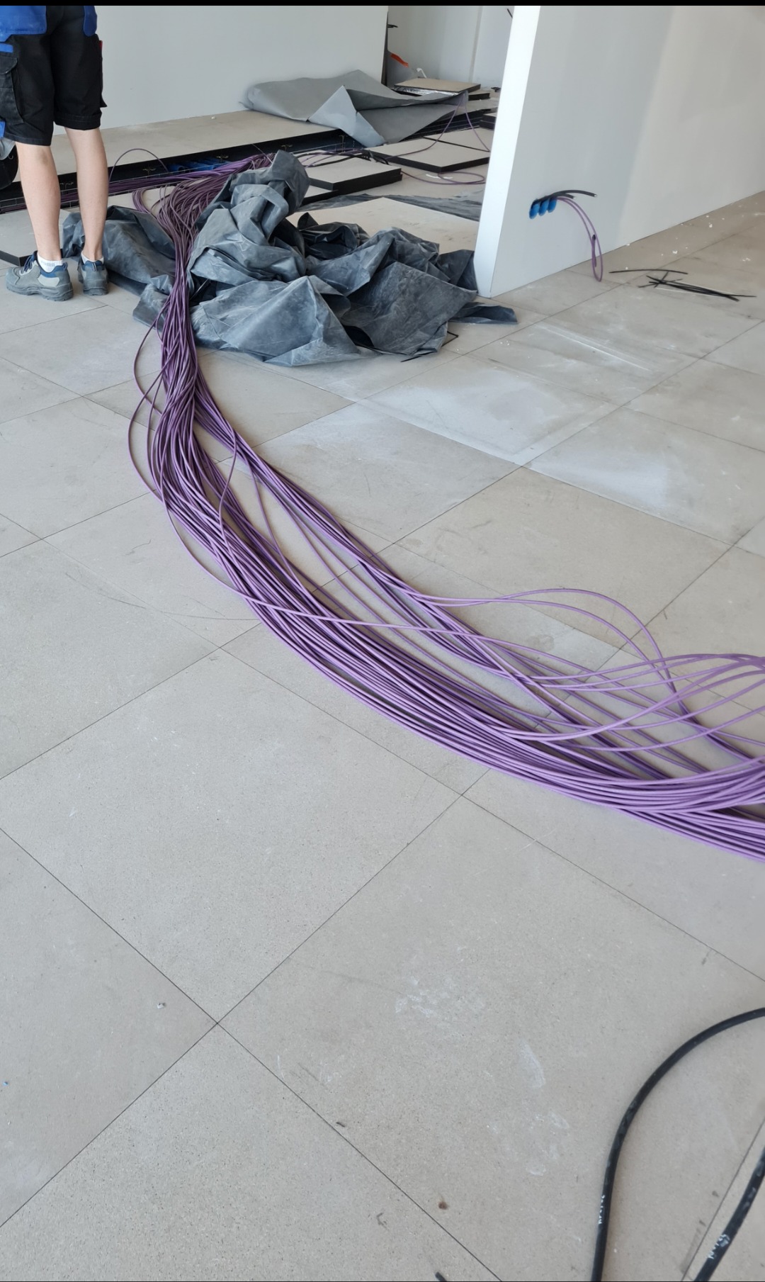 Fioletowe przewody elektryczne rozłożone na podłodze z płytek podczas instalacji, widoczne skrzynki elektryczne w ścianie i fragment instalatora w tle.