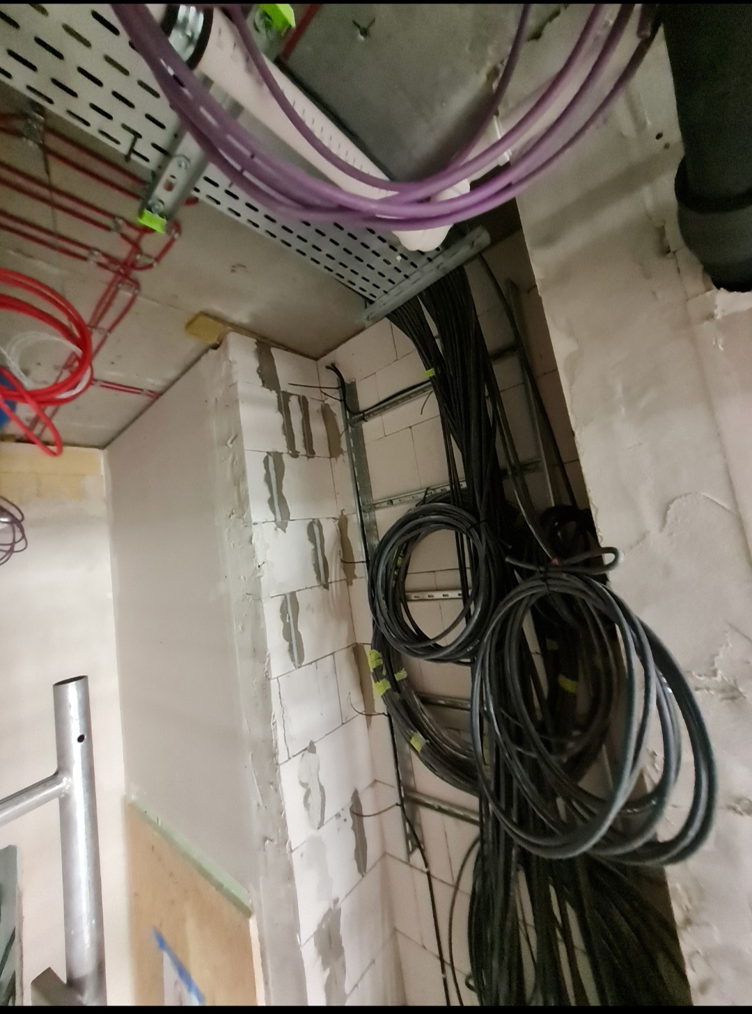Wnętrze w trakcie prac instalacyjnych: kable prowadzone w korytach i zwisające ze ściany z bloczków, widoczne fragmenty rusztowania i instalacji pod sufitem.