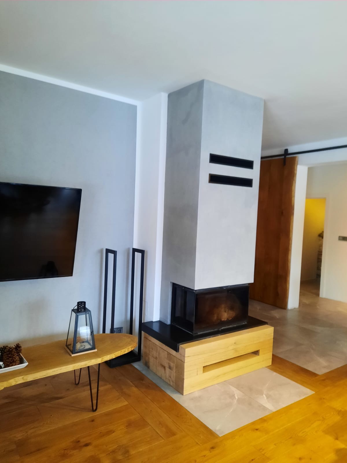 Nowoczesny salon z kominkiem obudowanym szarym betonem architektonicznym, drewnianym parkietem ułożonym we wzór jodełki francuskiej oraz przesuwnymi drzwiami z surowego drewna