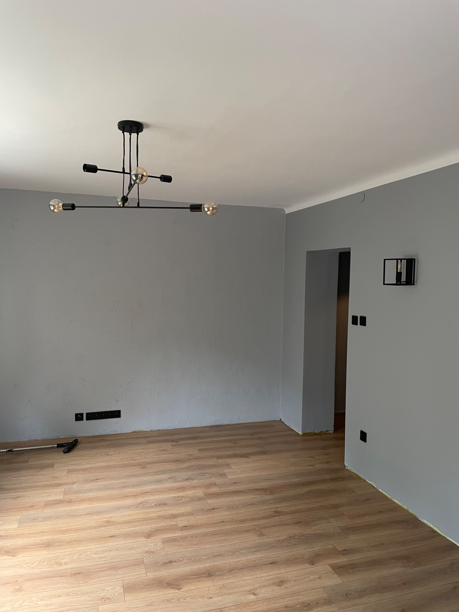 Wnętrze pokoju po remoncie: świeżo położone panele podłogowe w odcieniach brązu, szare ściany z widocznymi gniazdkami elektrycznymi i nowoczesnym oświetleniem sufitowym w stylu industrialnym.