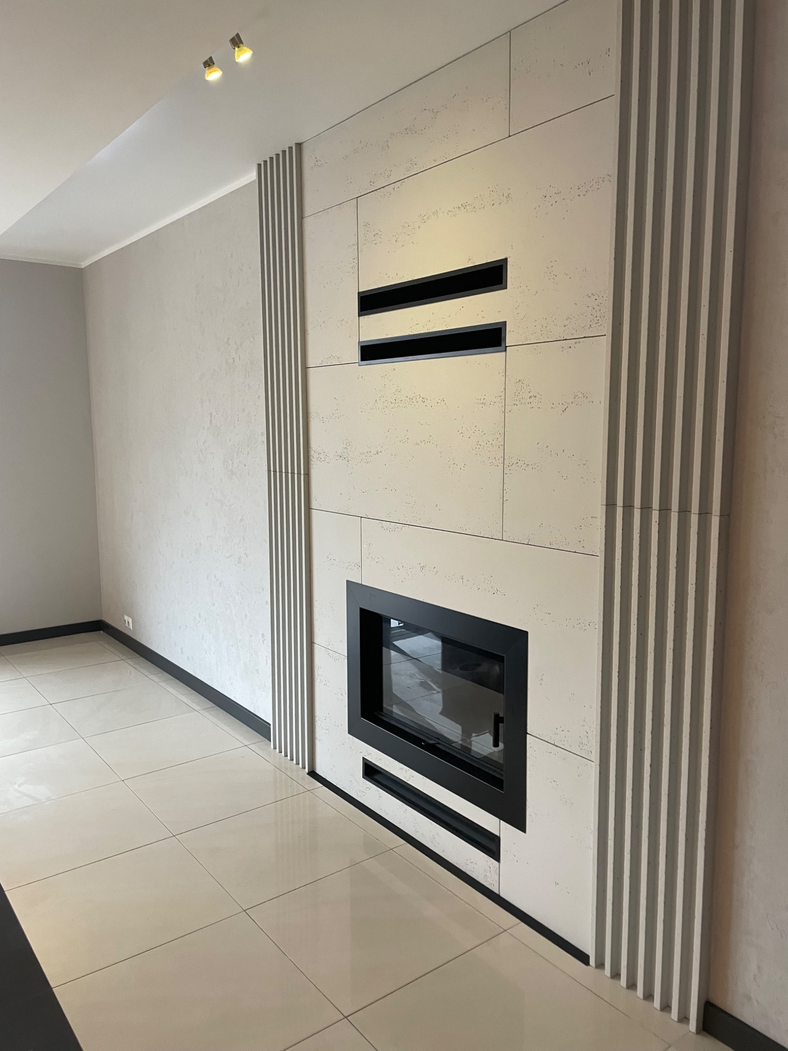 Nowoczesny salon z wykończonym kominkiem obłożonym płytami imitującymi trawertyn, z czarnym wkładem kominkowym i dekoracyjnymi pionowymi listwami po bokach, na podłodze jasne płytki.