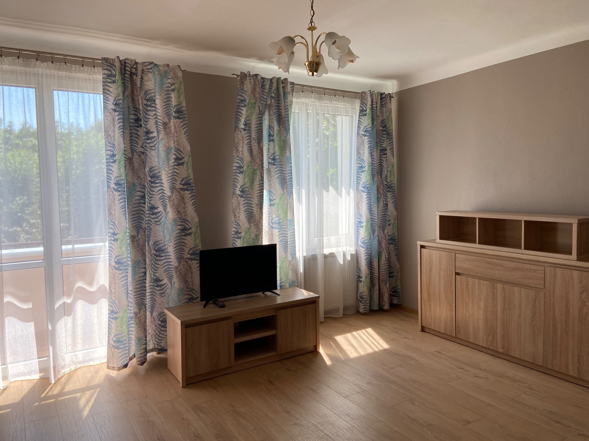 Jasne wnętrze pokoju z drewnianą podłogą, szafkami i telewizorem, okna zasłonięte wzorzystymi zasłonami w odcieniach zieleni i błękitu, widoczny fragment balkonu.