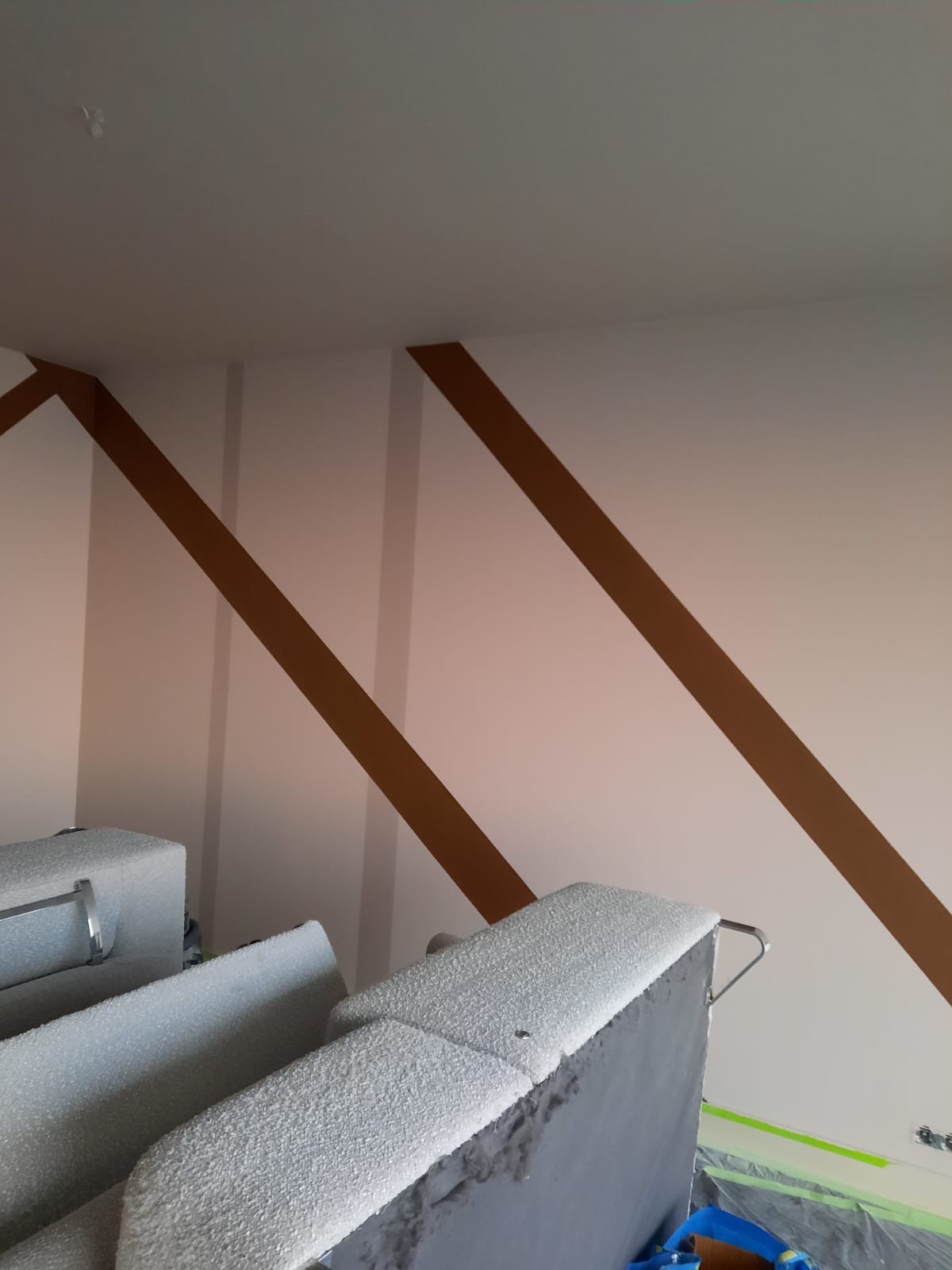 Ściana z świeżo pomalowanymi brązowymi pasami, zabezpieczona taśmą malarską, w tle zakryte folią meble.