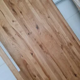 Fragment blatu z jasnego drewna dębowego z widocznymi sękami i naturalnym usłojeniem, ułożonego ukośnie na tle białej ściany.