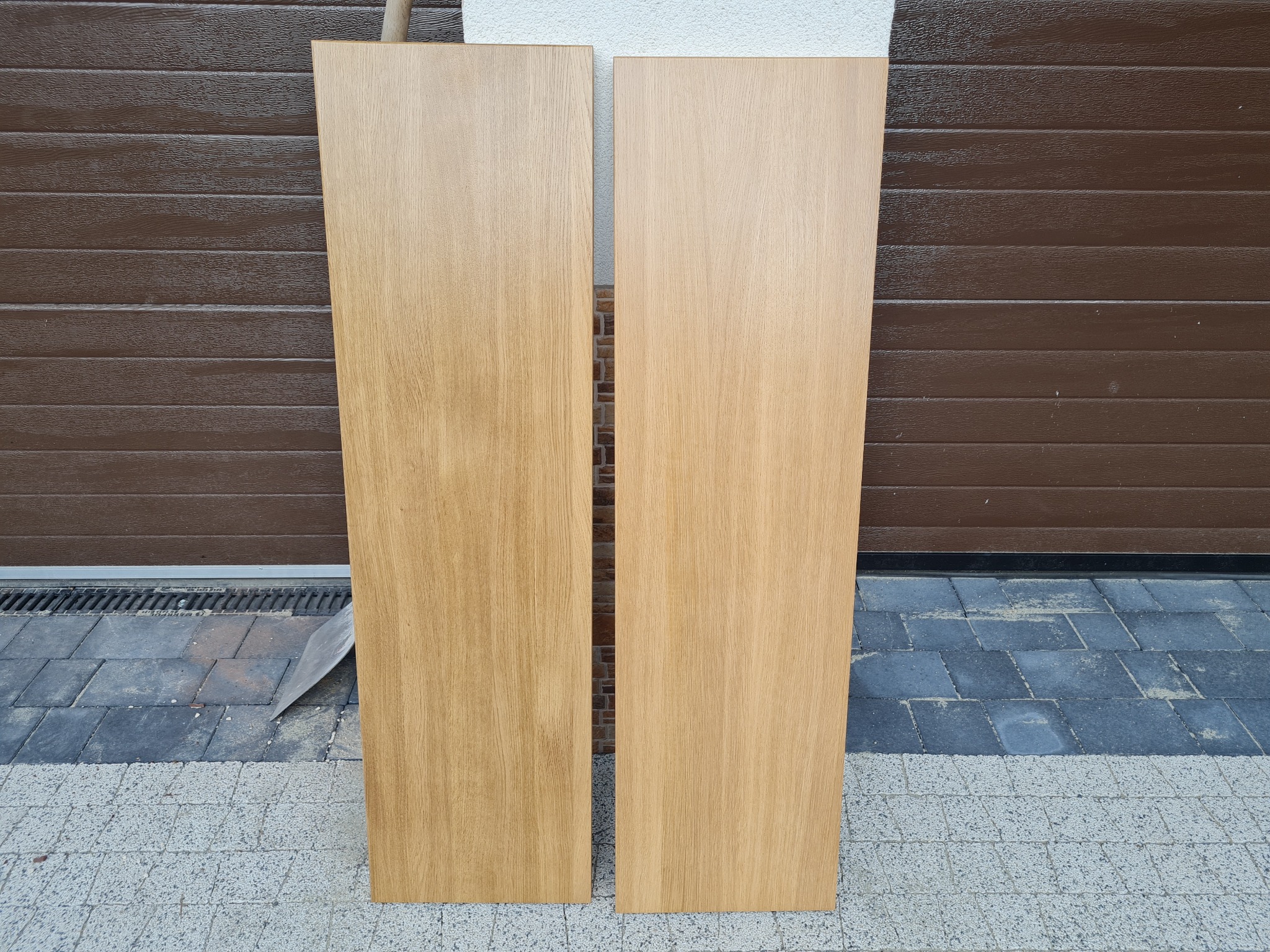 Dwa surowe, prostokątne elementy z jasnego drewna dębowego, o gładkiej powierzchni, oparte o brązową bramę garażową i kostkę brukową. Widoczna struktura drewna i naturalne odcienie.