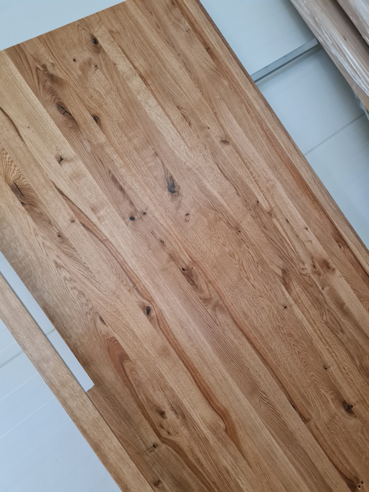 Fragment blatu z jasnego drewna dębowego z widocznymi sękami i naturalnym usłojeniem, ułożonego ukośnie na tle białej ściany.