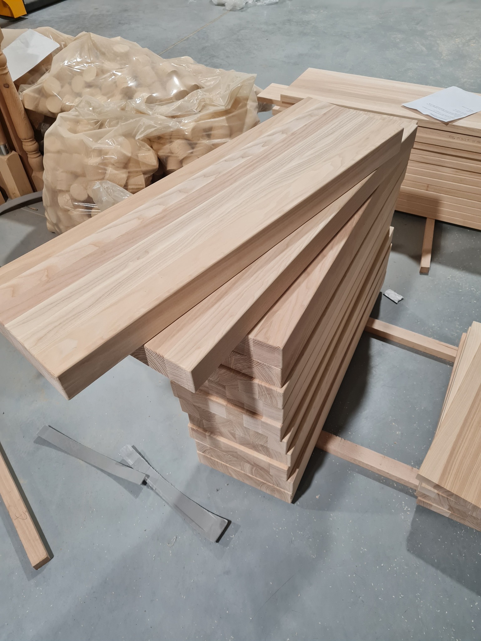 Stos surowych, jasnych desek drewnianych ułożonych na drewnianych legarach w hali produkcyjnej, obok worki z drewnianymi elementami i sterta desek.