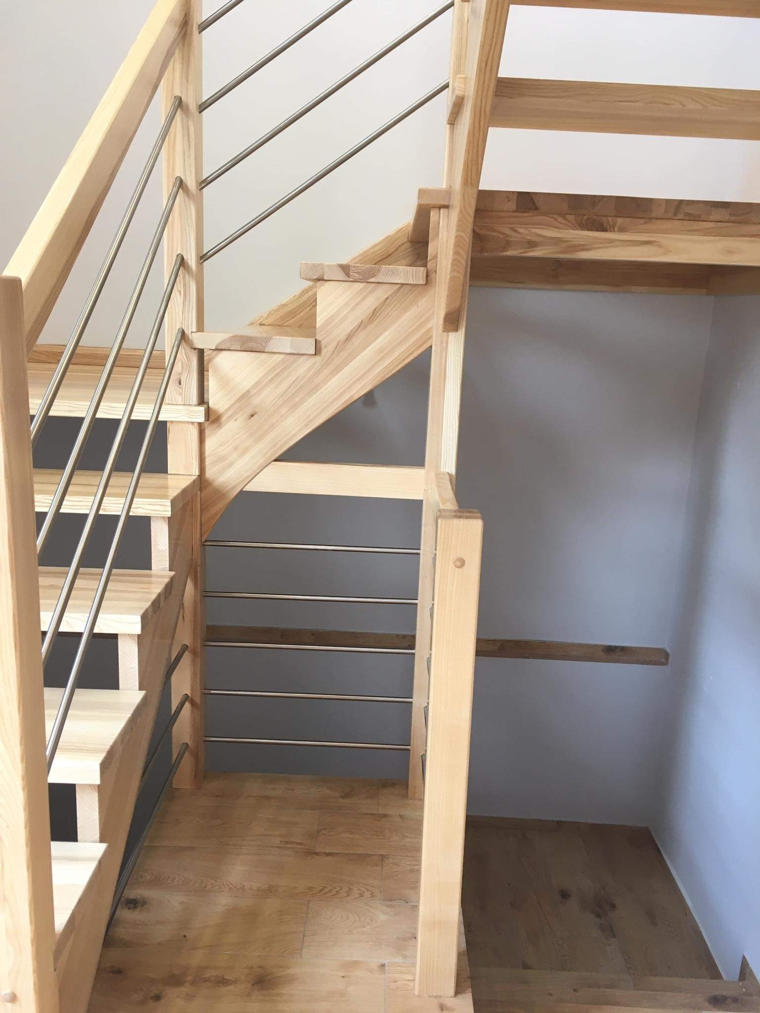 Jasne drewniane schody z metalowymi balustradami, widoczne sęki na drewnie i szara ściana w tle.