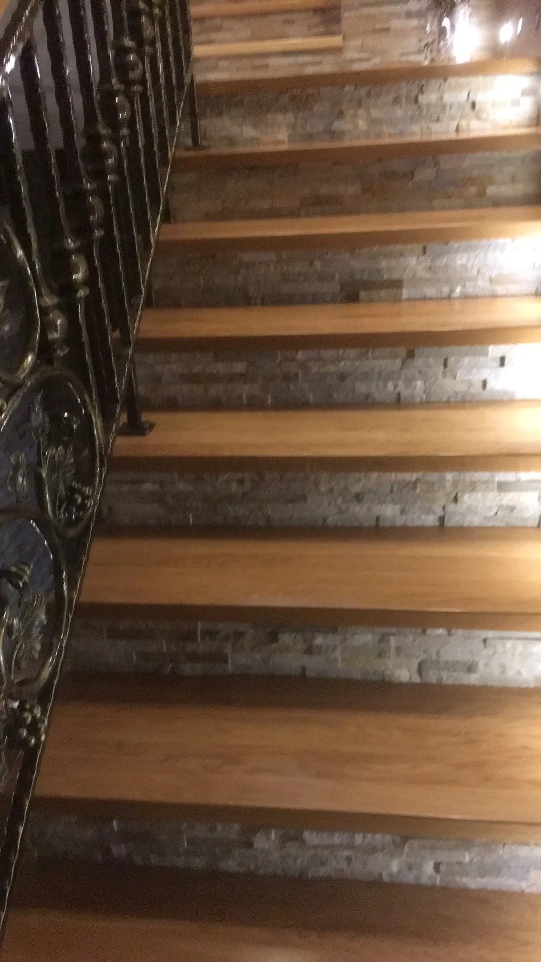 Klatka schodowa z drewnianymi stopniami i dekoracyjną, metalową balustradą z motywami roślinnymi na tle ściany wyłożonej kamieniem dekoracyjnym.