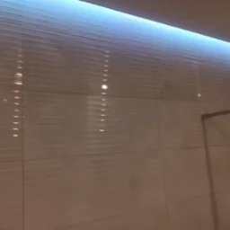 Ściana łazienki wyłożona białymi, poziomymi płytkami z subtelnym wzorem, oświetlona od góry niebieskim światłem LED.