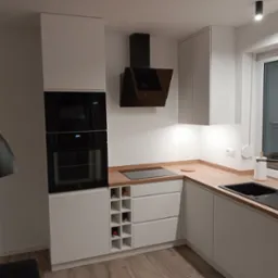 Nowoczesna kuchnia w odcieniach bieli i drewna z czarnymi akcentami: okap, zlew i lampa wisząca. Zabudowa kuchenna z piekarnikiem, mikrofalówką, płytą indukcyjną i wbudowanym regałem na wino.