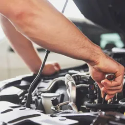 Mężczyzna w Strzałkowie, w niebieskim kombinezonie, naprawia silnik samochodu za pomocą klucza nasadowego. Widoczny otwarty przód auta i fragment białego samochodu w tle.