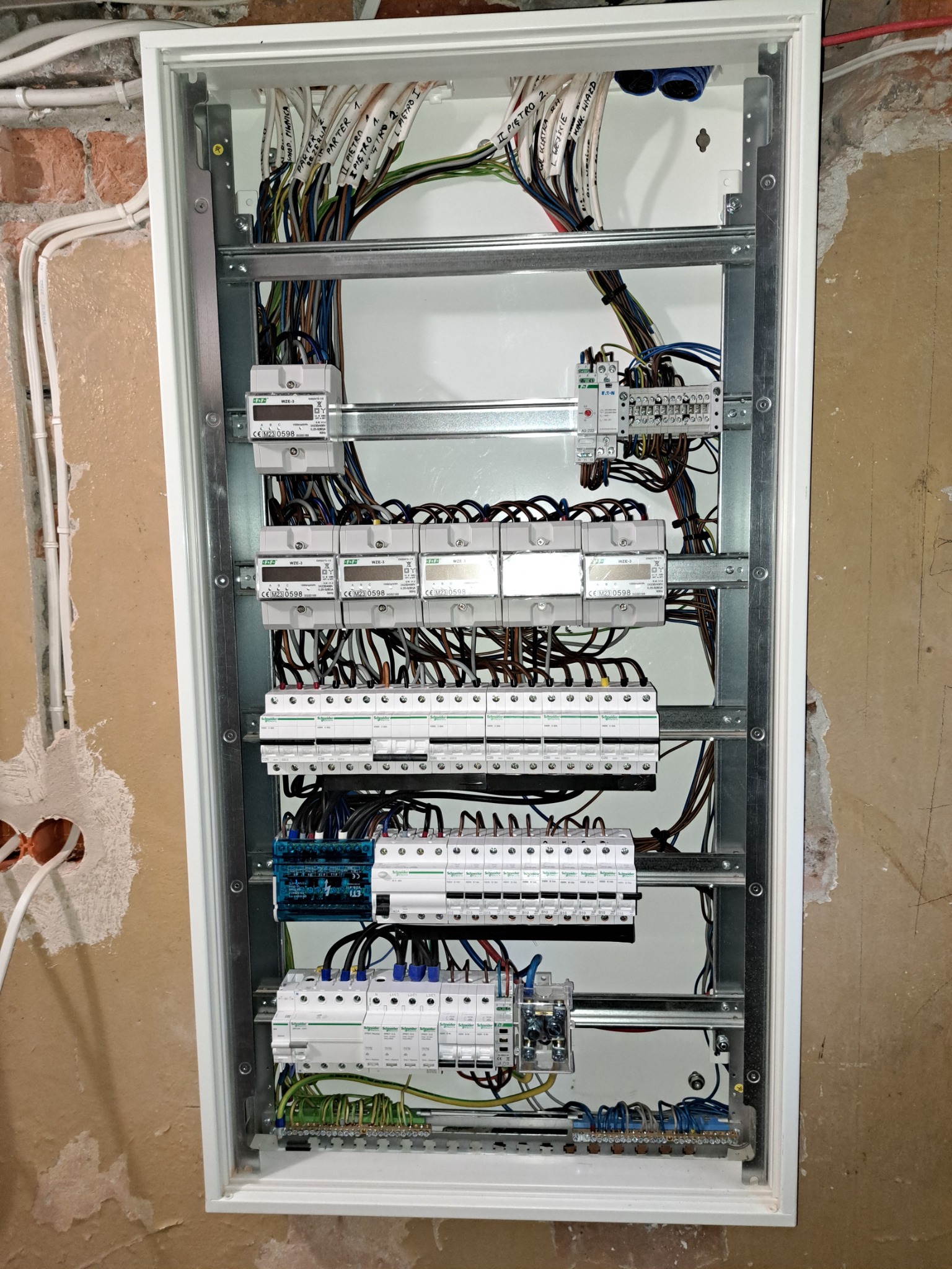 Otwarta skrzynka rozdzielcza z licznikami i zabezpieczeniami, widoczne przewody opisane etykietami, montaż w surowym stanie, nieotynkowana ściana w tle.