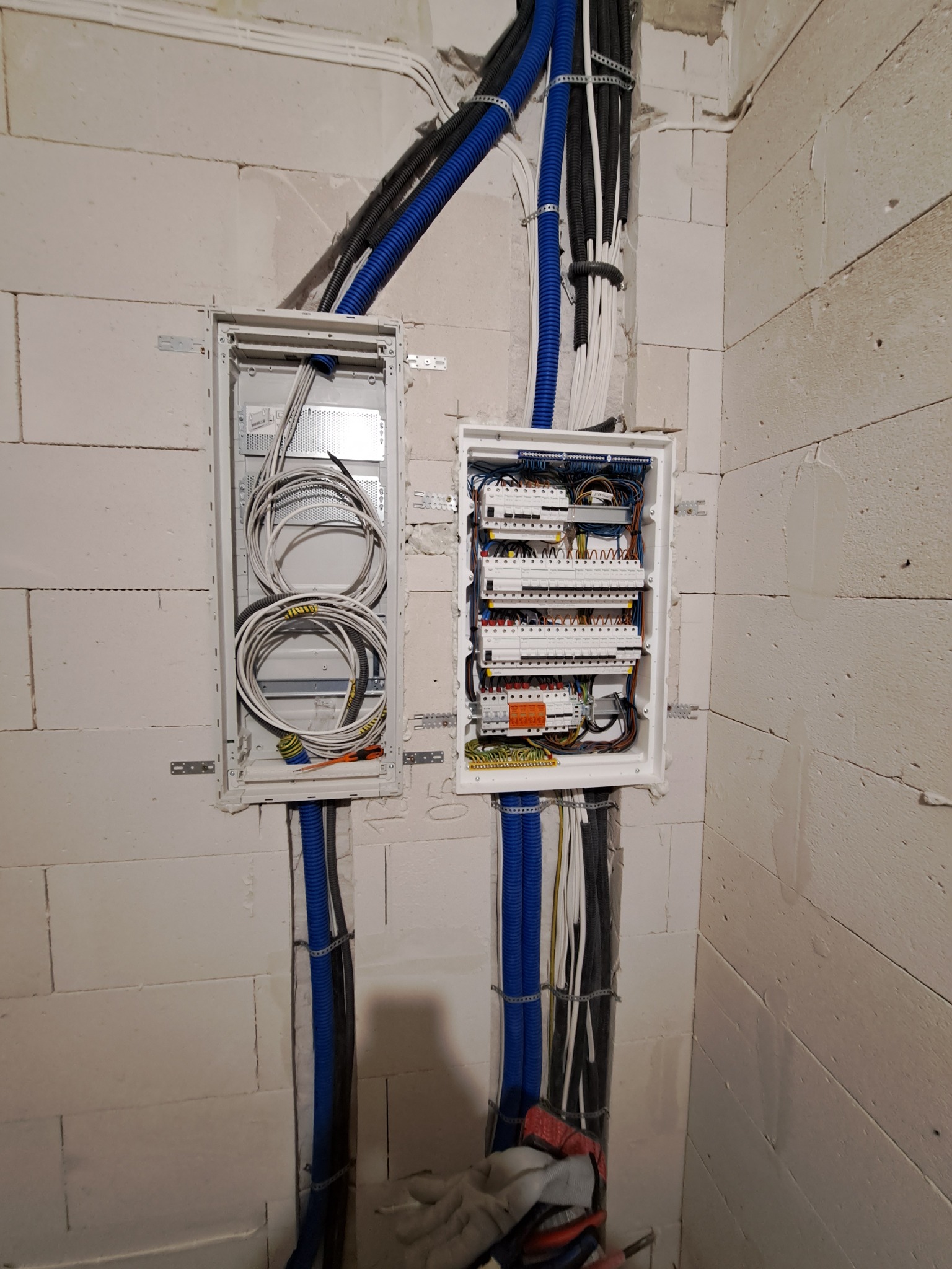 Dwa otwarte panele elektryczne zamontowane na ścianie z białych pustaków, z widocznymi przewodami w peszlach, jeden panel z luźno ułożonymi kablami, drugi z podłączonymi bezpiecznikami i elementami...