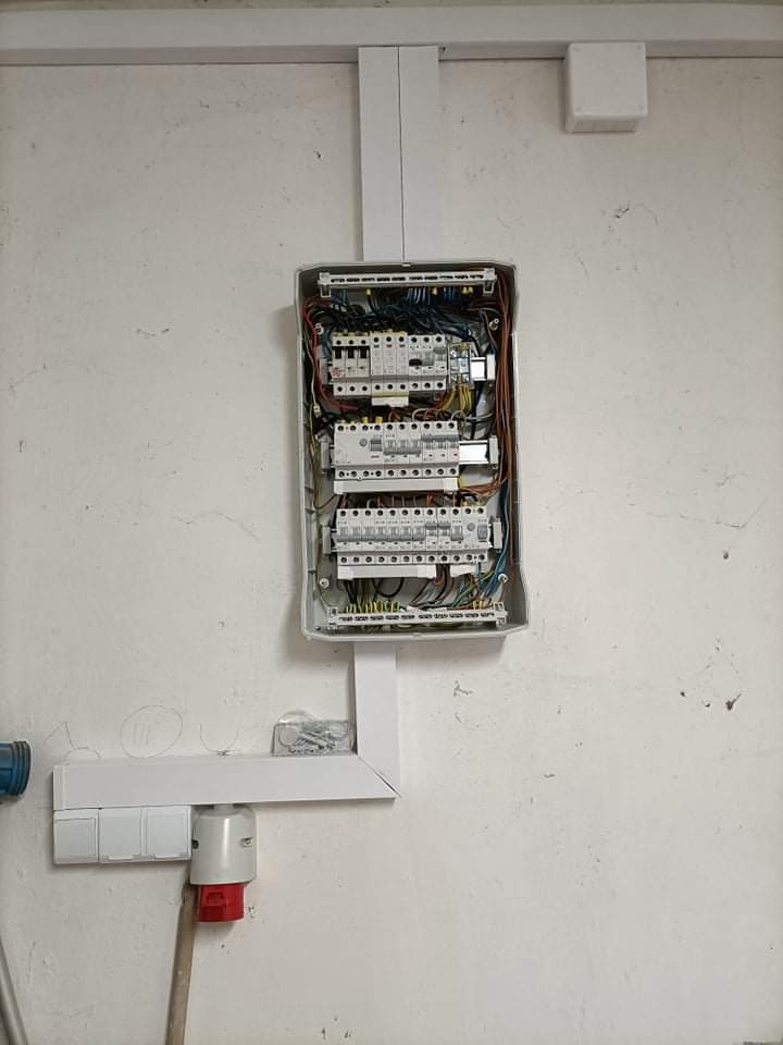 Otwarta skrzynka elektryczna z widocznymi bezpiecznikami i przewodami, zamontowana na szorstkiej, białej ścianie. Obok widoczny panel z wyłącznikami i gniazdem siłowym.