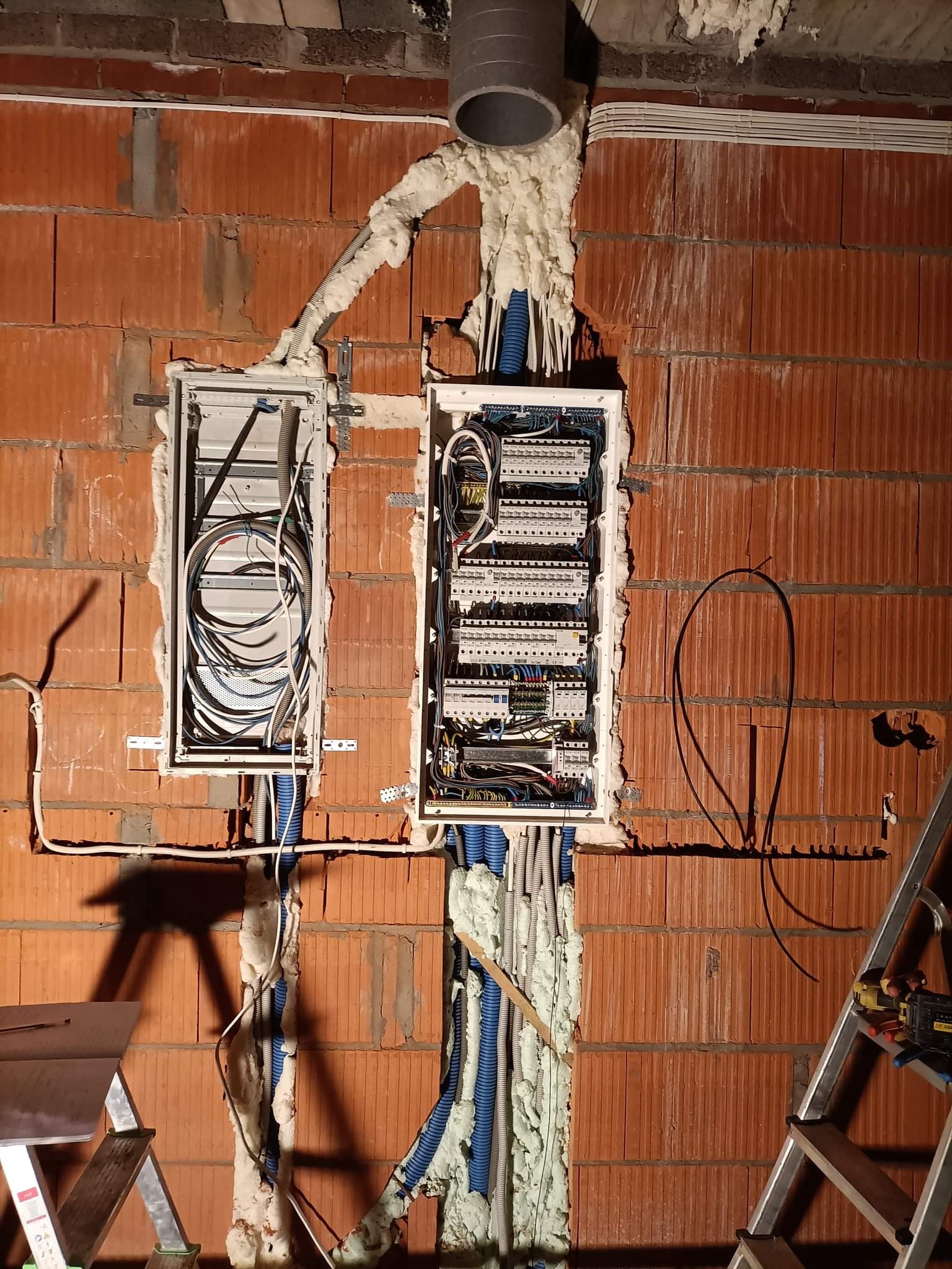 Dwa otwarte panele elektryczne w trakcie montażu na ścianie z cegły, zabezpieczone pianką montażową, widoczne kable i przewody, drabina w tle.