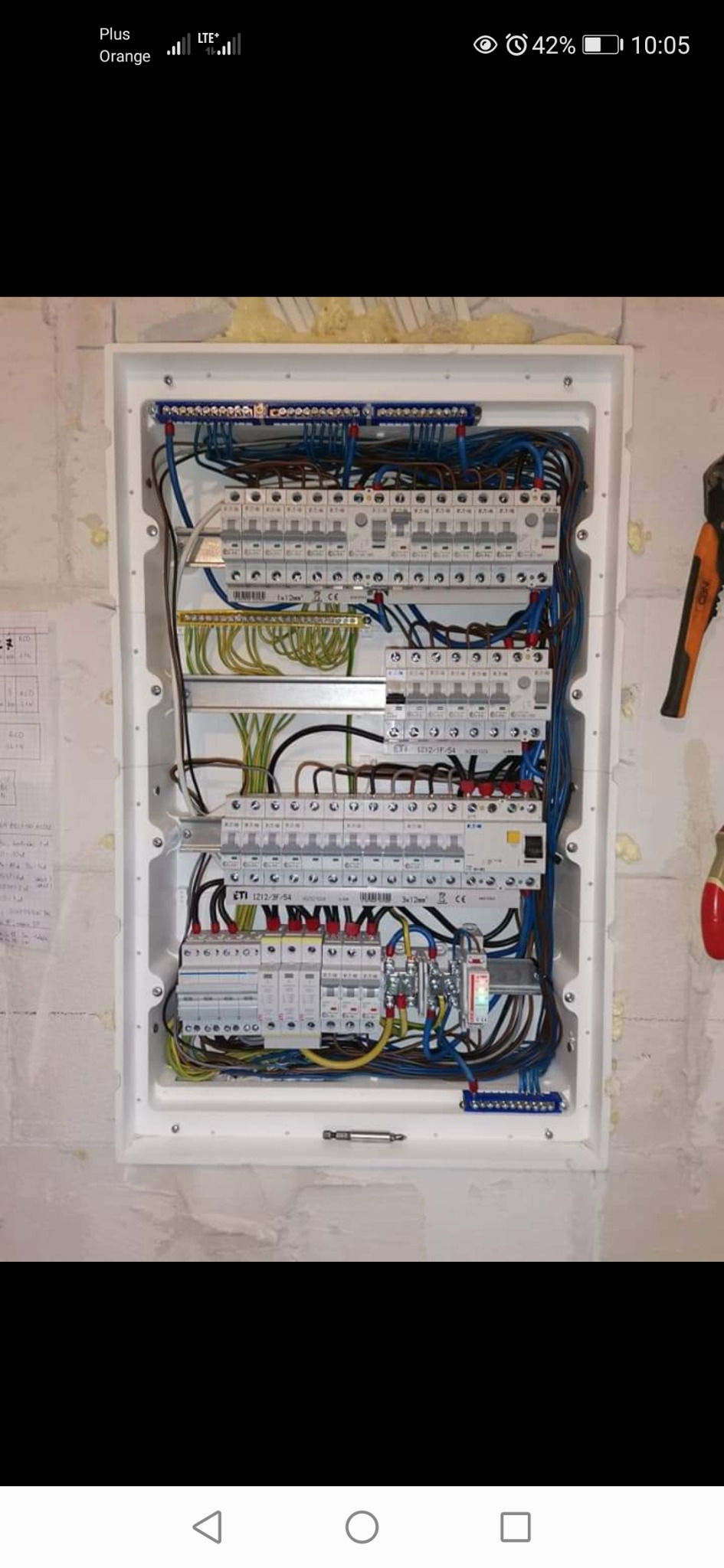 Rozdzielnica elektryczna z zainstalowanymi bezpiecznikami i okablowaniem w białej obudowie, zamontowana na surowej ścianie. Widoczne narzędzia obok.