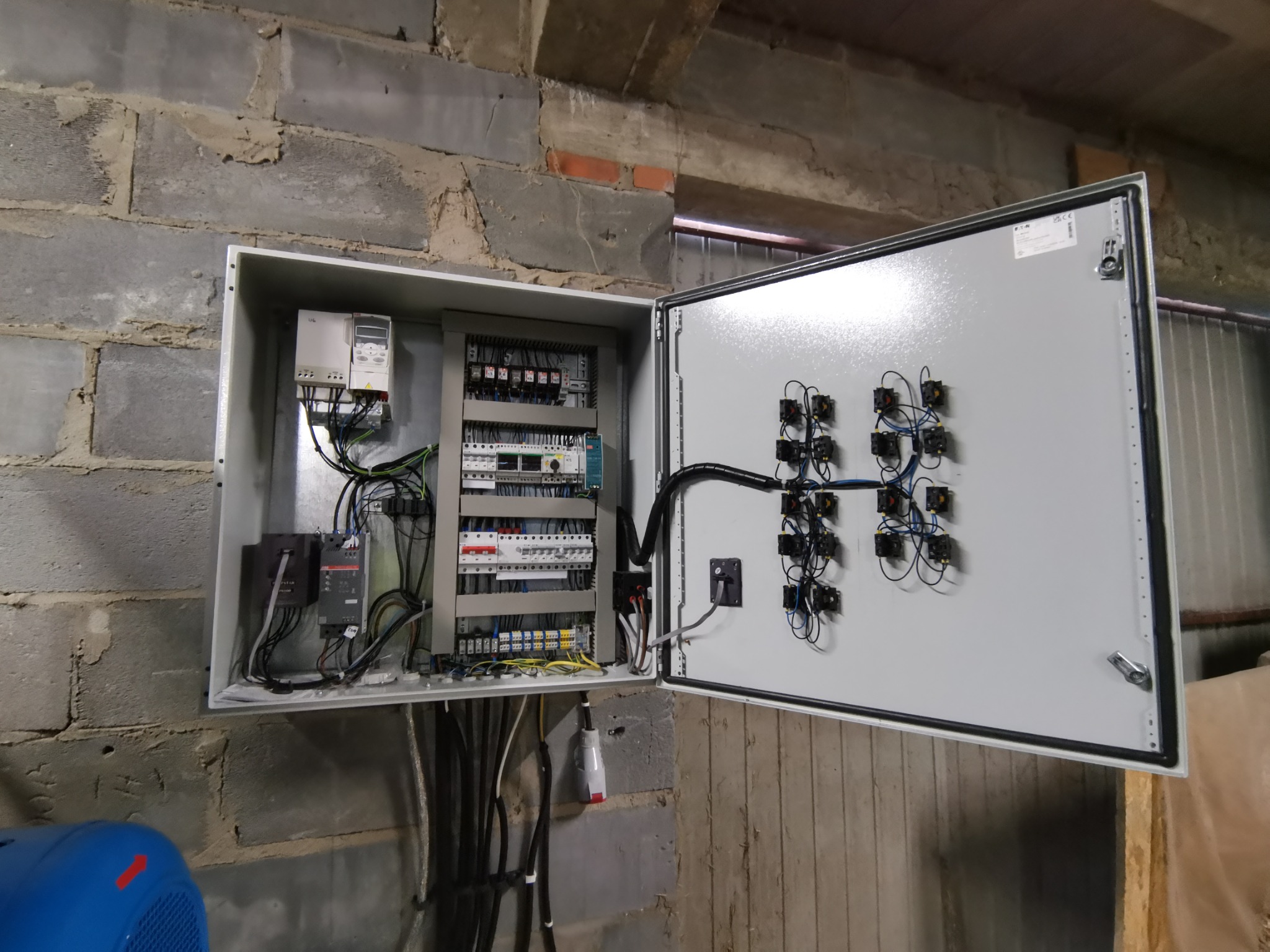 Otwarta szafa sterownicza z widocznymi elementami elektrycznymi, przekaźnikami i okablowaniem, zamontowana na surowej ścianie z cegły.