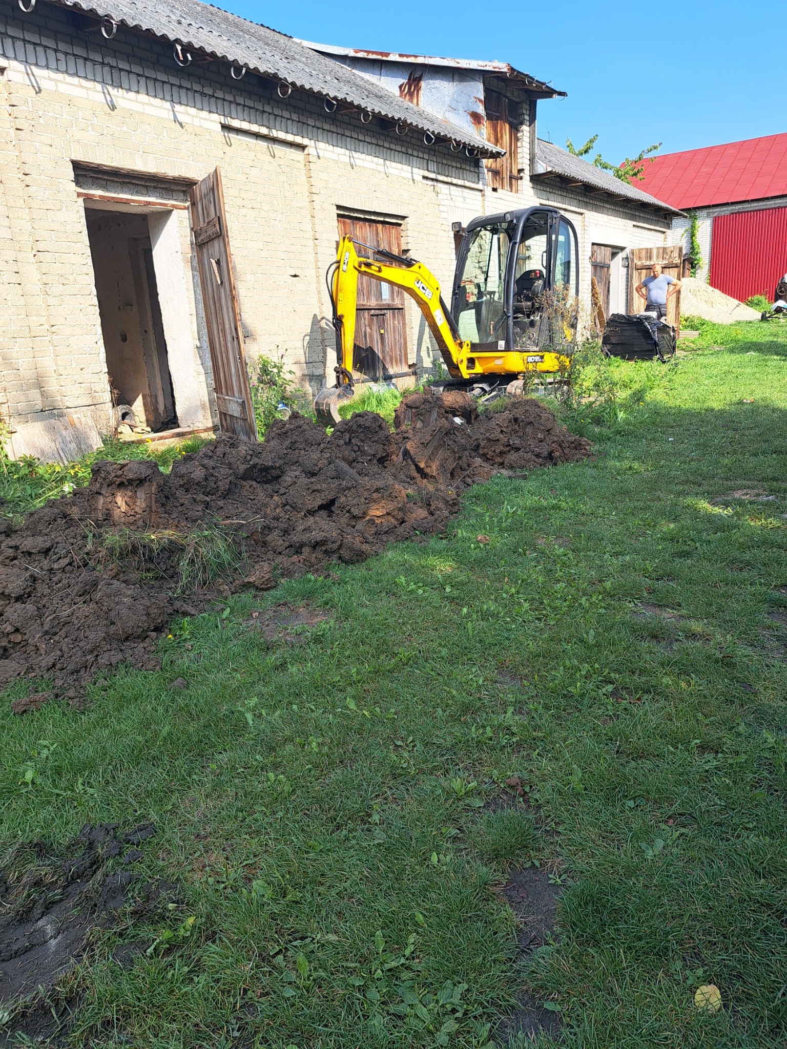 Żółta mini koparka JCB na trawniku przed budynkiem z cegły, świeżo wykopany rów z brudną ziemią, mężczyzna w tle.