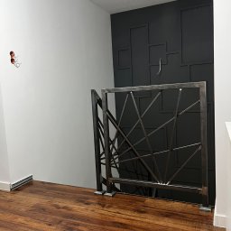 Weld Service - Metalowa balustrada schodowa o geometrycznym wzorze na tle ściany z dekoracyjnymi panelami w odcieniach szarości i brązu, widok z dołu na drewnianą podłogę.