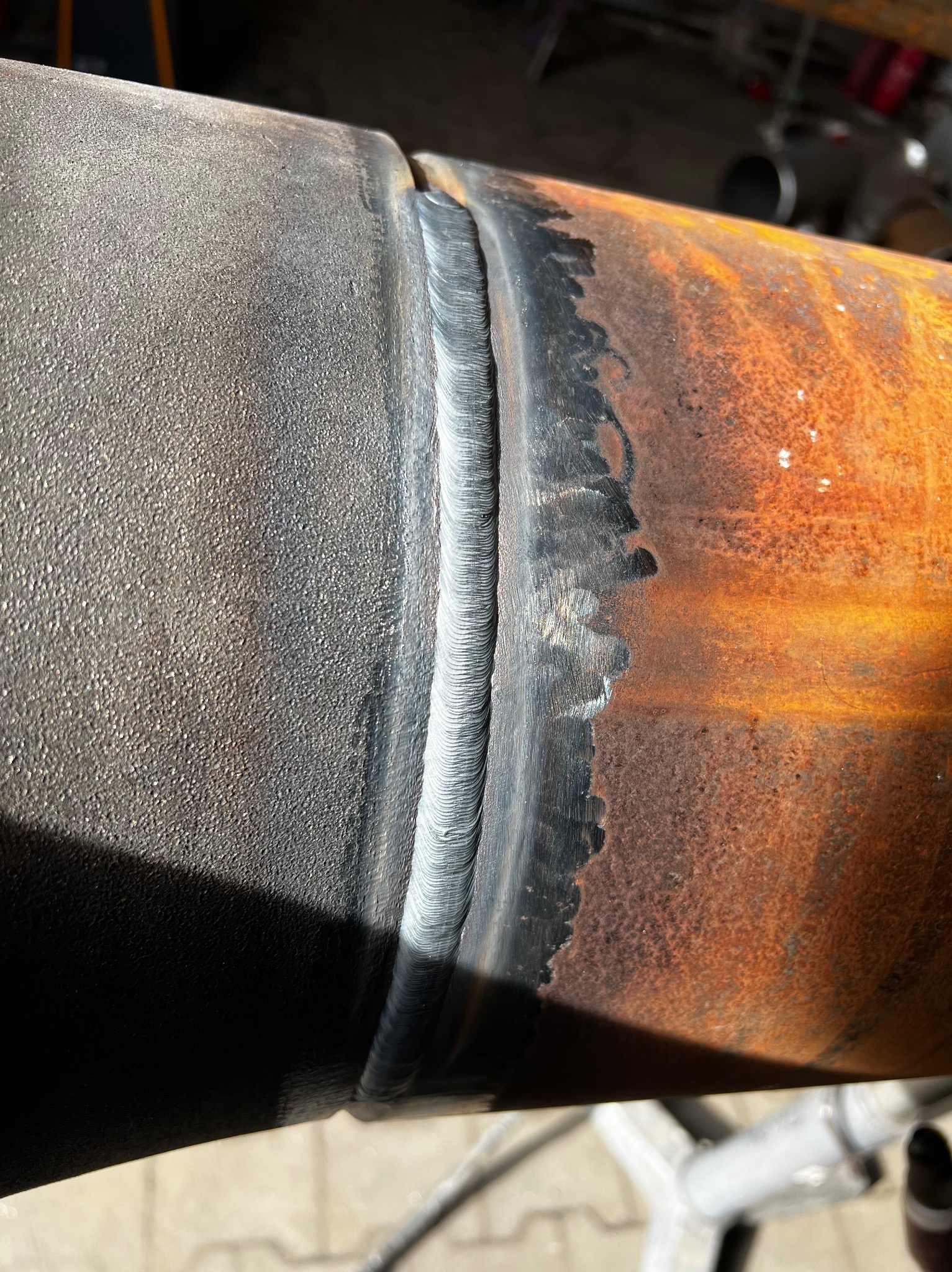 Spoina na rurze o zróżnicowanej powierzchni, od szorstkiej i ciemnej do rdzewiejącej, z widocznymi śladami po spawaniu.