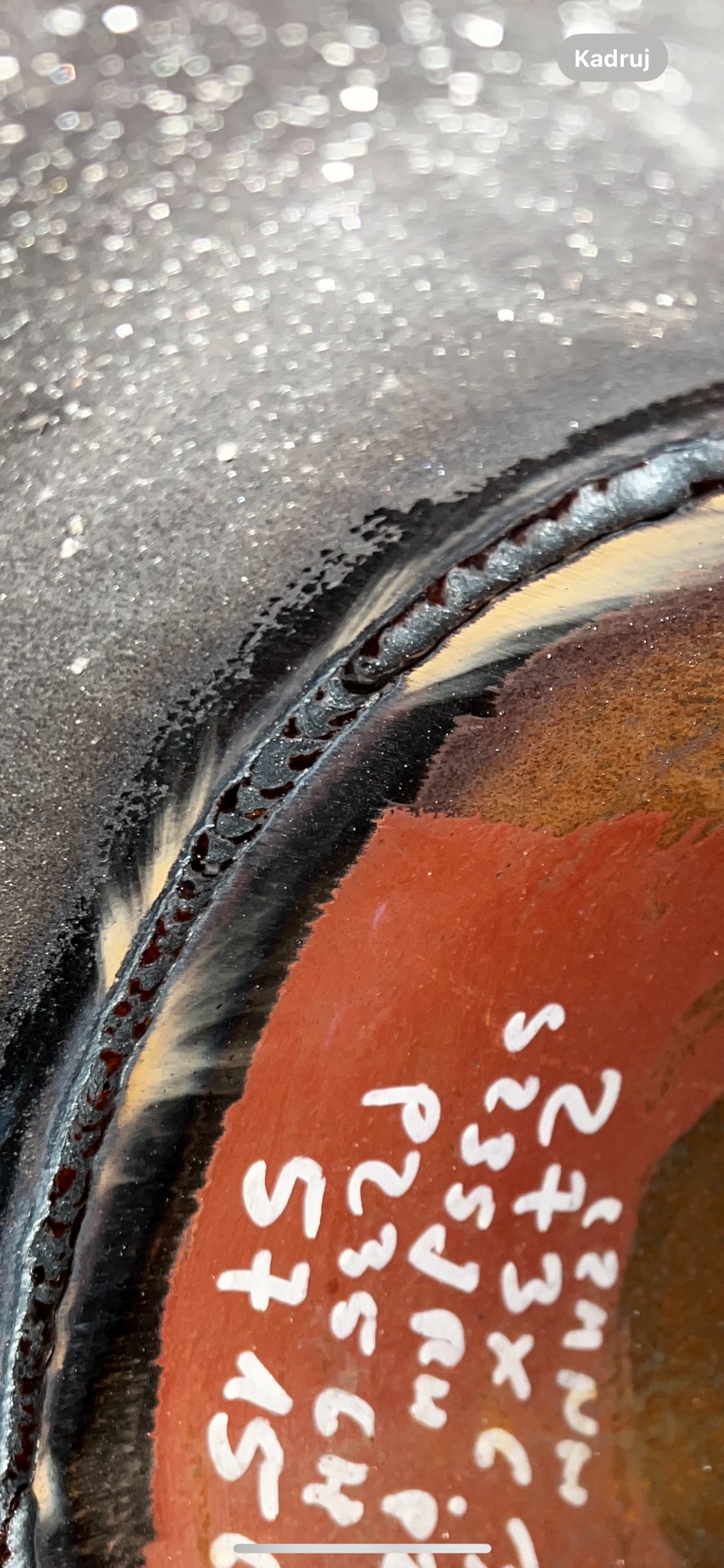 Zbliżenie na spoinę na metalowej rurze z oznaczeniami naniesionymi białą farbą, widoczne drobne nierówności i struktura spoiny.