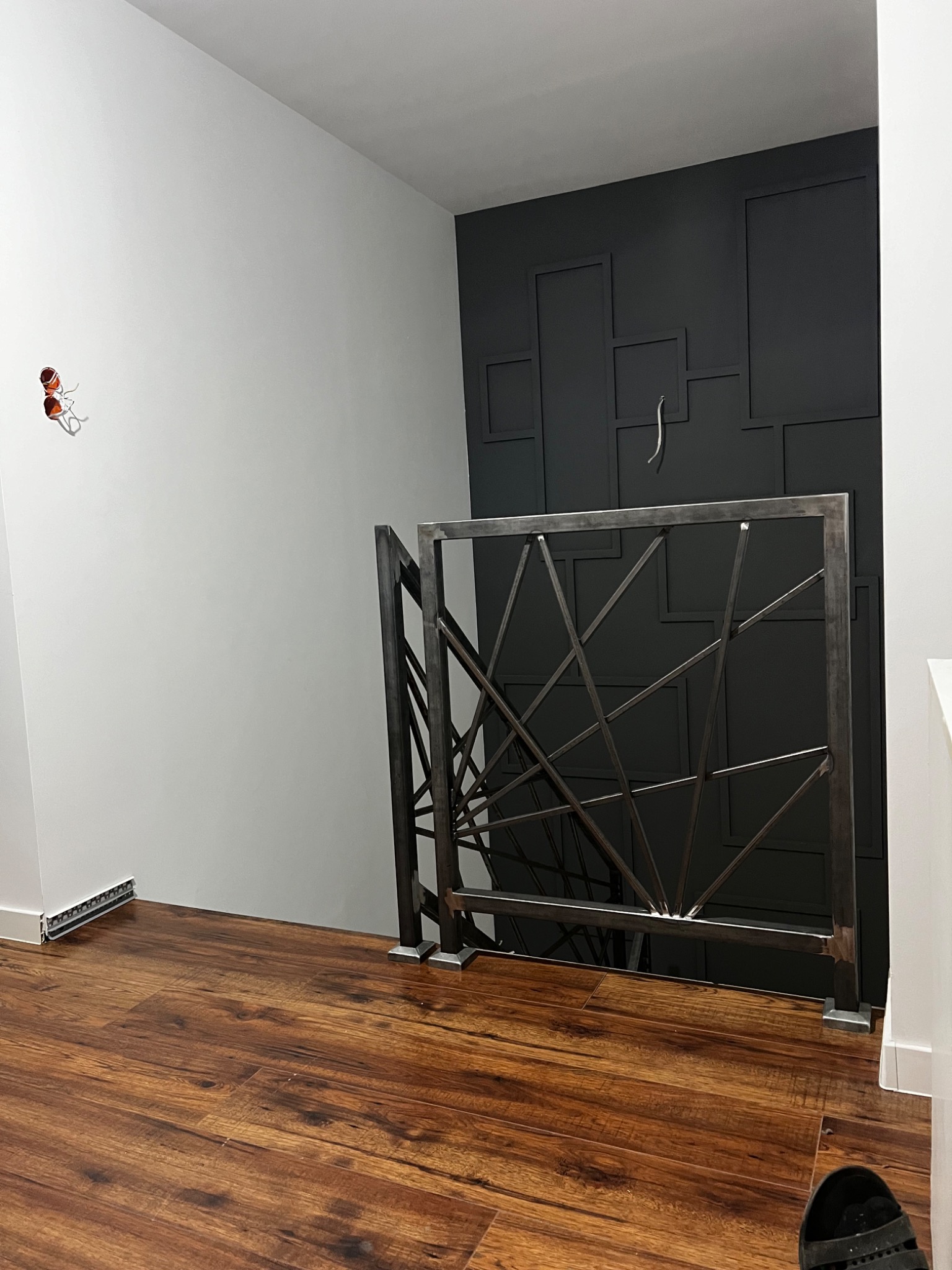 Metalowa balustrada schodowa o geometrycznym wzorze na tle ściany z dekoracyjnymi panelami w odcieniach szarości i brązu, widok z dołu na drewnianą podłogę.