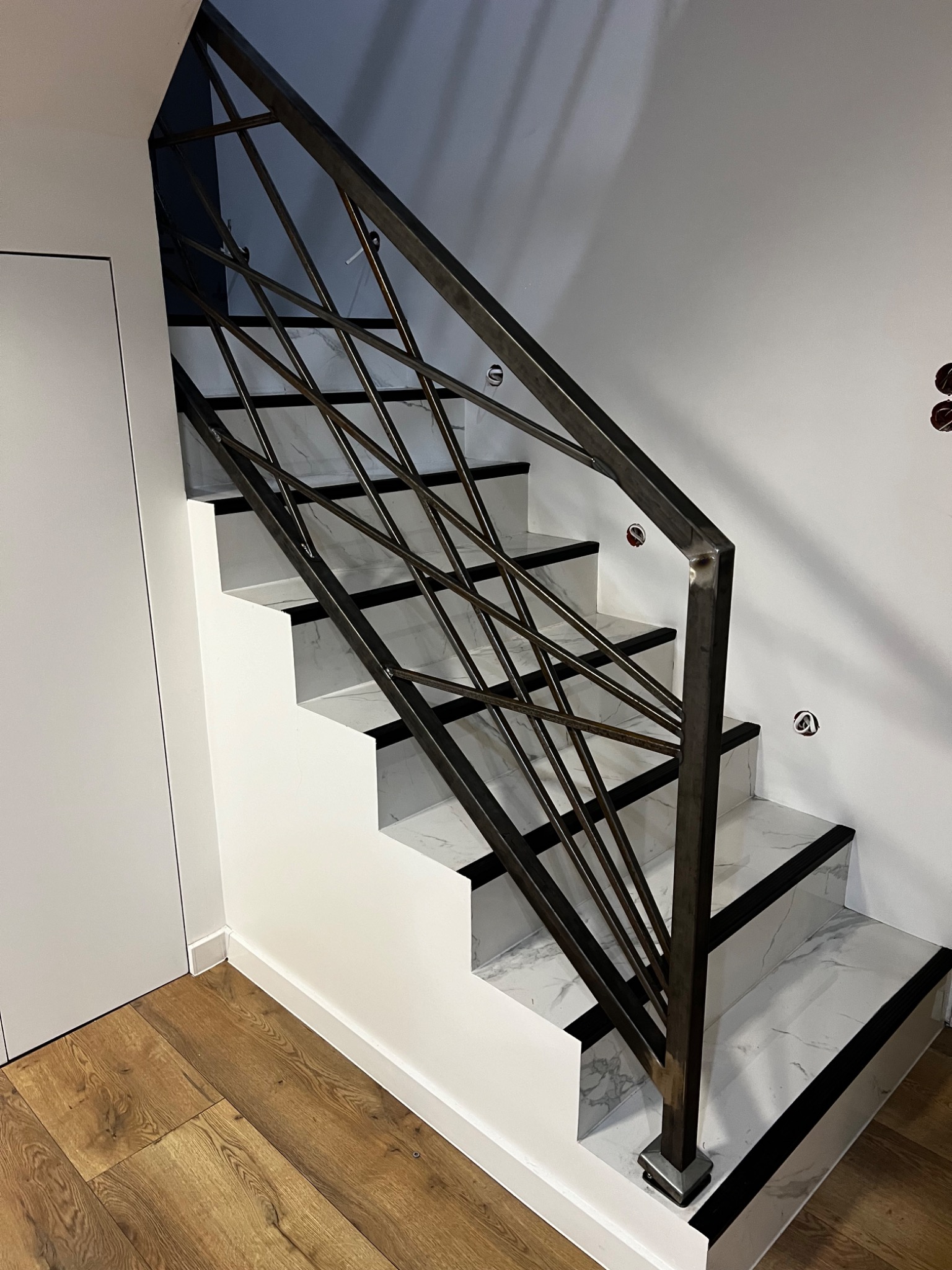 Nowoczesne schody z metalową balustradą o geometrycznym wzorze, wykończone białym marmurem z czarnymi akcentami, na tle drewnianej podłogi i białych ścian.