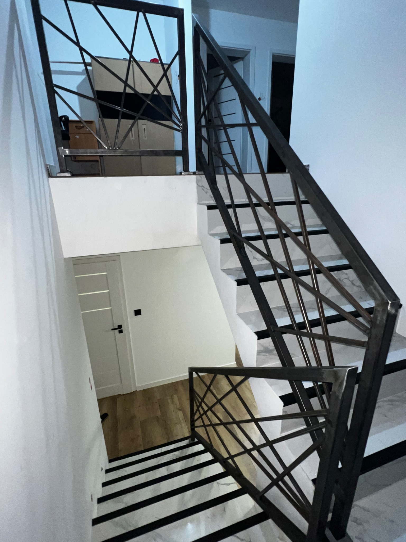 Nowoczesne schody z metalową balustradą o geometrycznym wzorze, białe ściany i marmurowe stopnie z czarnymi paskami, widok z góry.