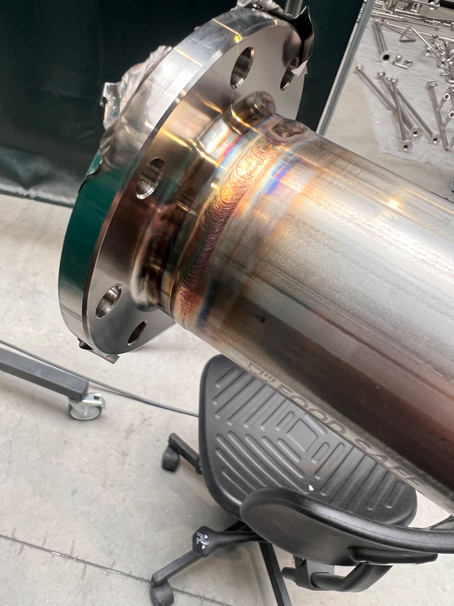 Błyszczący, spawany element z metalu nierdzewnego z kołnierzem, widoczne ślady po spawaniu i przebarwienia termiczne, leżący na krześle biurowym.