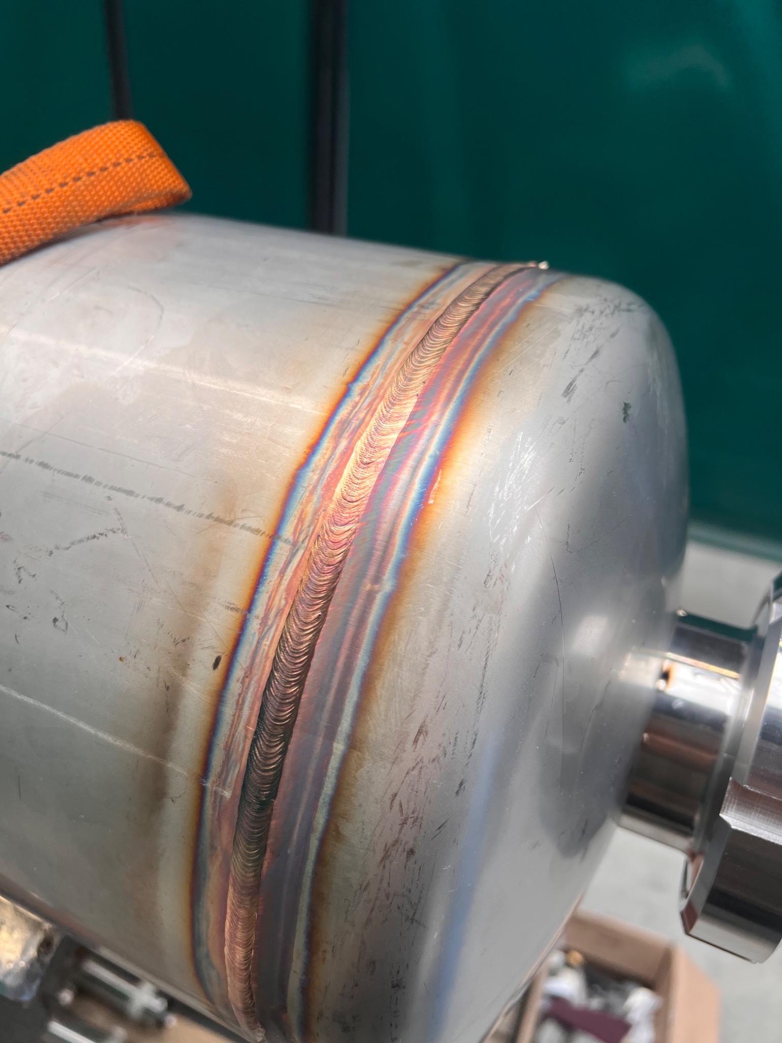 Zbliżenie na spoinę na metalowym zbiorniku, widoczne tęczowe przebarwienia po spawaniu, zarys pomarańczowego pasa transportowego w górnej części.