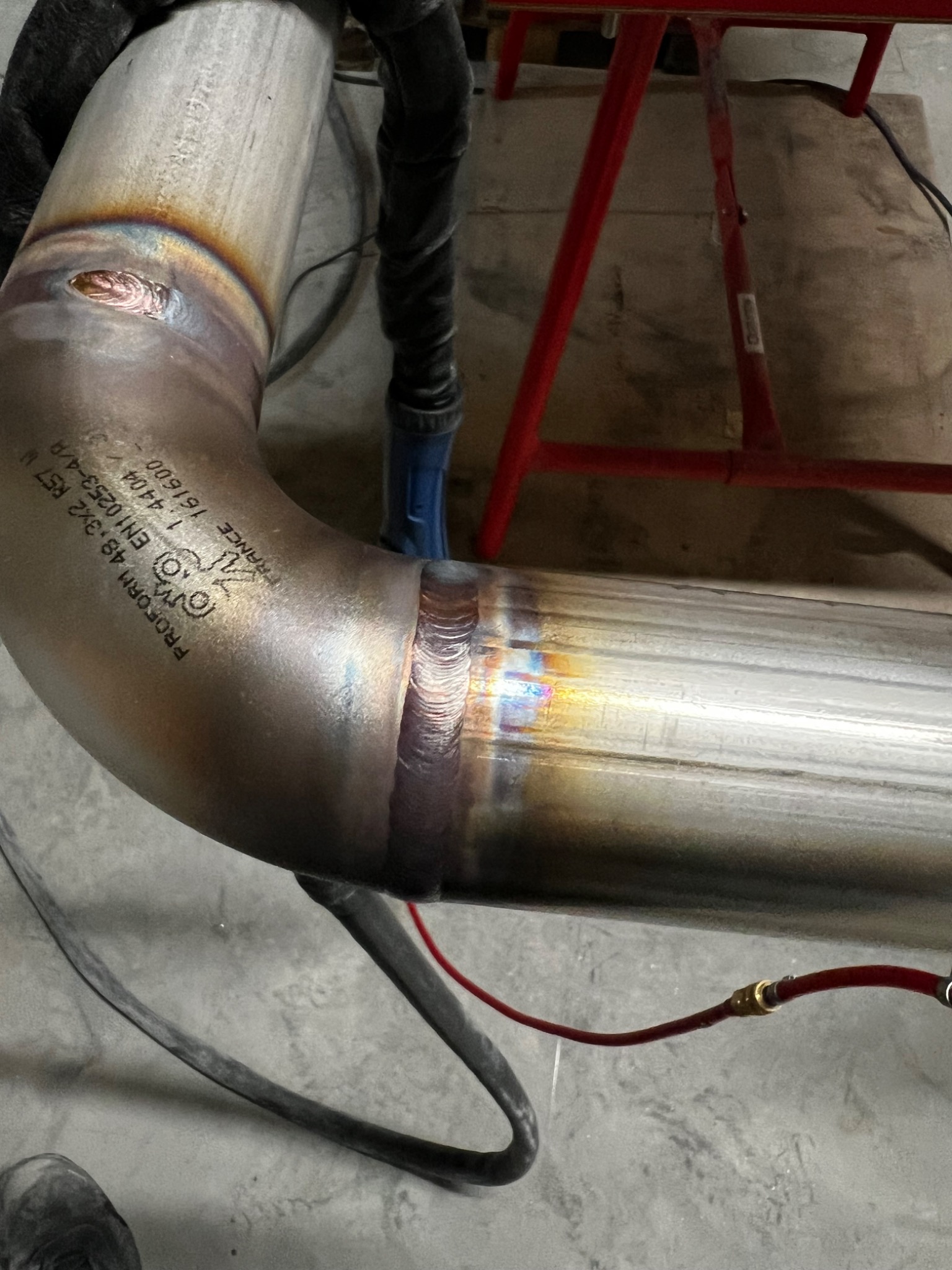 Spoina na zagiętej rurze ze stali nierdzewnej, widoczne ślady termiczne po spawaniu i oznaczenia producenta rury, ustawiona na czerwonym stojaku.