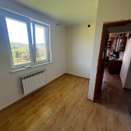 Dariusz Kulig usługi Remontowo-Budowlane - Jasny pokój po malowaniu i położeniu gładzi: drewniana podłoga, białe ściany, okno z widokiem na zieleń, grzejnik. Widoczne przejście do kuchni.