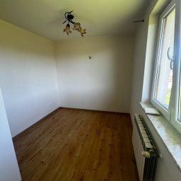 Dariusz Kulig usługi Remontowo-Budowlane - Jasne wnętrze po malowaniu: drewniana podłoga, białe ściany z grzejnikiem pod oknem. Widoczna lampa sufitowa. Świeżo wykończone pomieszczenie.