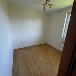 Dariusz Kulig usługi Remontowo-Budowlane - Jasne wnętrze pokoju po malowaniu ścian i położeniu paneli podłogowych. Widoczny grzejnik, okno i lampa sufitowa. Ukończone wykończenie.