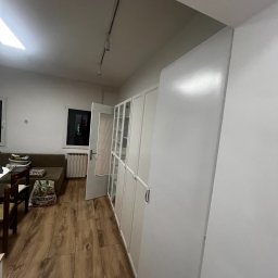 Dariusz Kulig usługi Remontowo-Budowlane - Białe szafy wnękowe z przeszklonymi drzwiami, widok na drewnianą podłogę i okno. Minimalistyczne wnętrze po remoncie, oświetlenie punktowe.