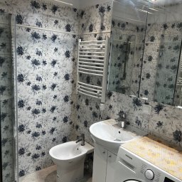 Dariusz Kulig usługi Remontowo-Budowlane - Wykończona łazienka z kabiną prysznicową, bidetem, umywalką z szafką i grzejnikiem drabinkowym. Ściany wyłożone płytkami w kwiatowy wzór, pralka pod blatem.